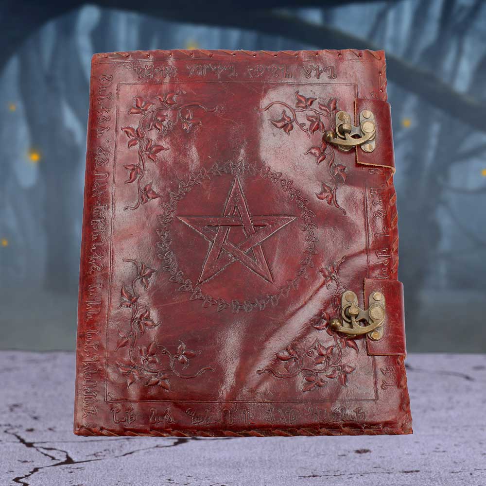 A red, leather-bound journal sits closed with two metal clasps. An embossed pentagram within a circular design is centered on the front, adorned with floral patterns. An indiscernible text surrounds the design's border. The journal is in a blurred outdoor setting.