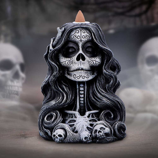 Sculpted female skull bust with closed eye sockets and decorative facial designs, topped by an incense cone, framed by flowing hair and small skulls against a foggy graveyard background.